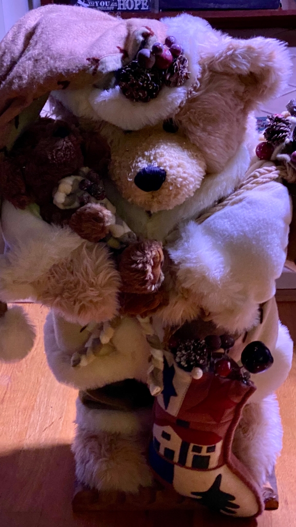 Large cream-colored stuffed teddy bear dressed in festive holiday garb holding a stuffed brown bear cub. 