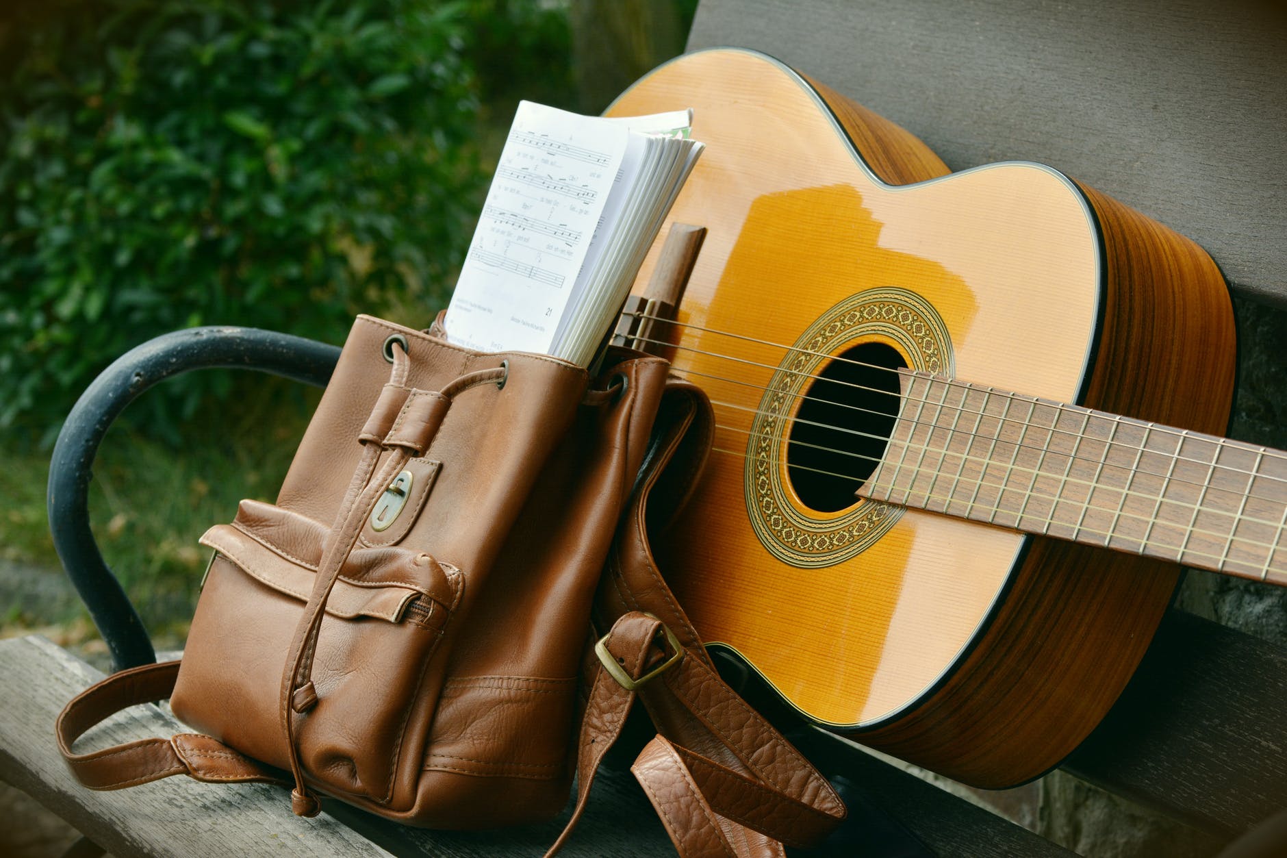 an acoustic guitar on a park bench with a leather satchel containing music
